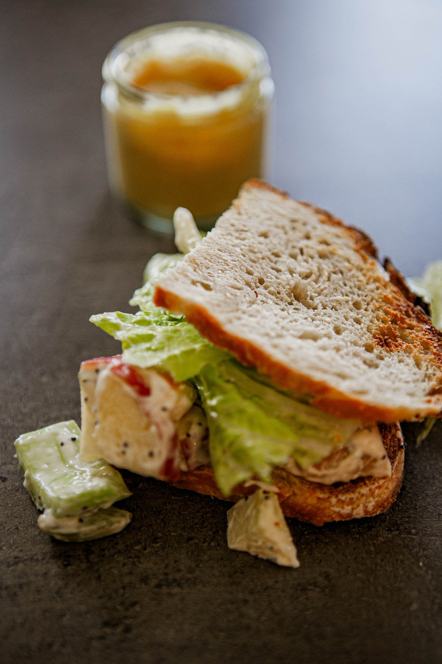 A chicken salad sandwich with lettuce on sourdough bread with a honey mustard dipping sauce on a dark surface. Made in the kitchen of Nystrom Orchard in Worthington Minnesota.