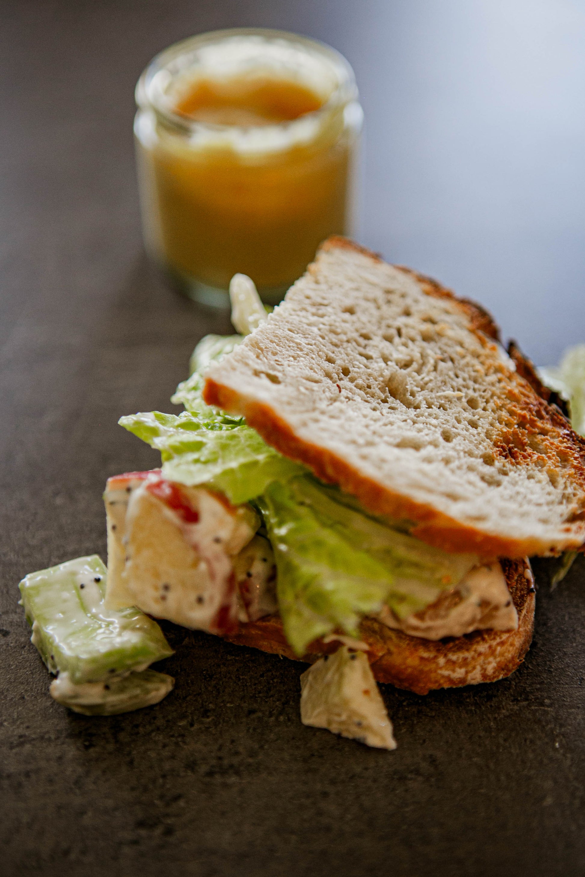 A chicken salad sandwich with lettuce on sourdough bread with a honey mustard dipping sauce on a dark surface. Made in the kitchen of Nystrom Orchard in Worthington Minnesota.