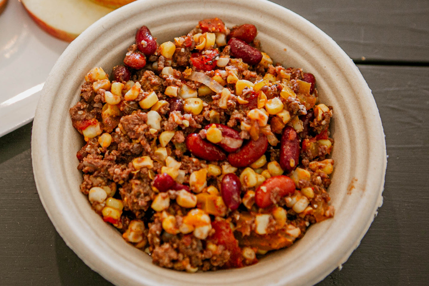 A bowl of hearty chili with visible ground beef, beans, corn and tomatoes on a wooden surface. Made in the kitchen of Nystrom Orchard in Worthington Minnesota.
