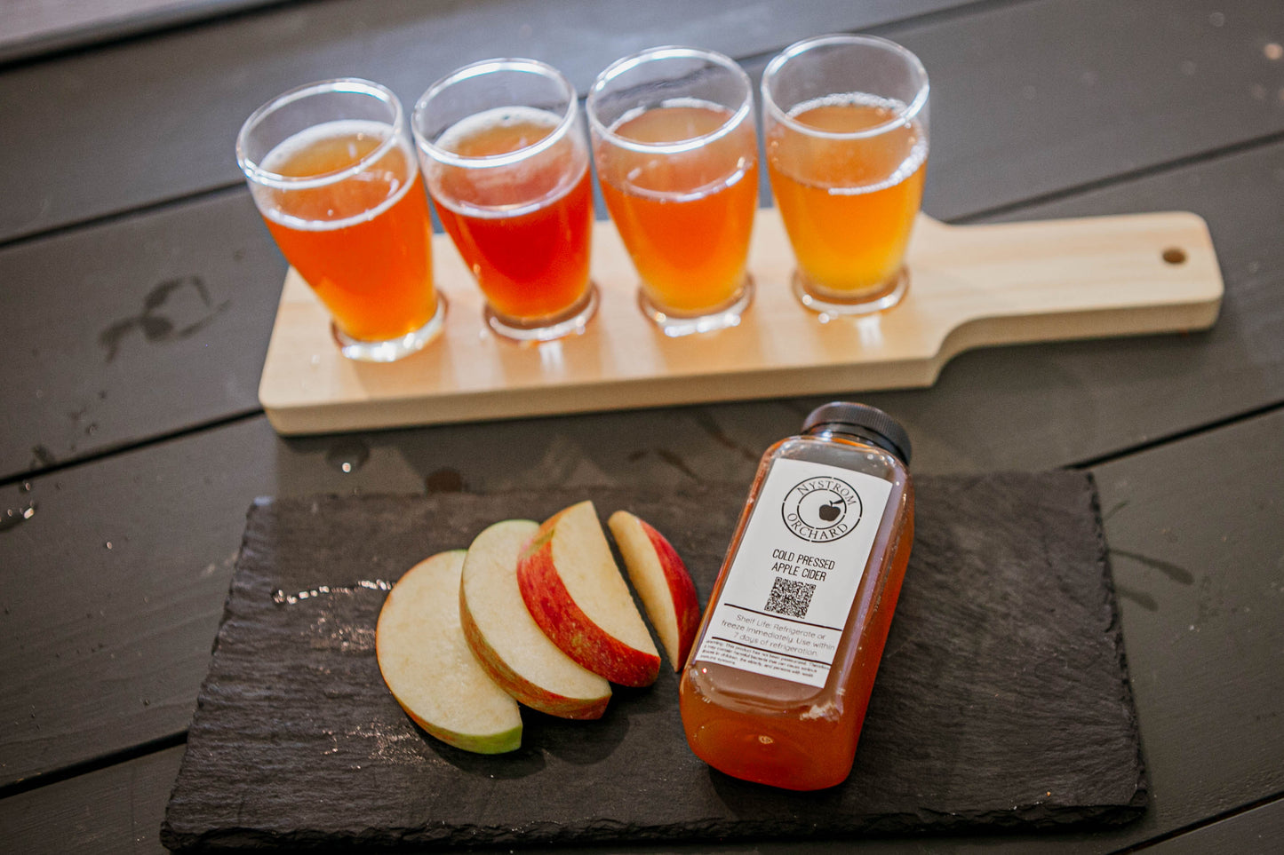 Cider flight on a board with sliced apples and a mini cold pressed cider on a dark surface. Pressed in the kitchen at Nystrom Orchard in Worthington Minnesota.