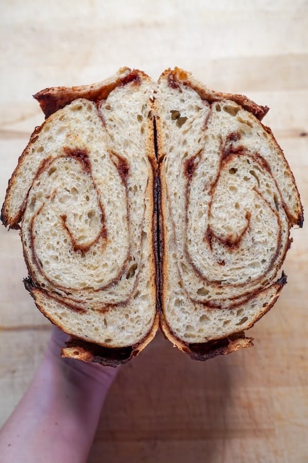 A hand holding a sliced loaf of rustic swirled cinnamon sourdough bread, showing the layered interior, against a light wooden background. Baked in the kitchen of Nystrom Orchard in Worthington Minnesota.
