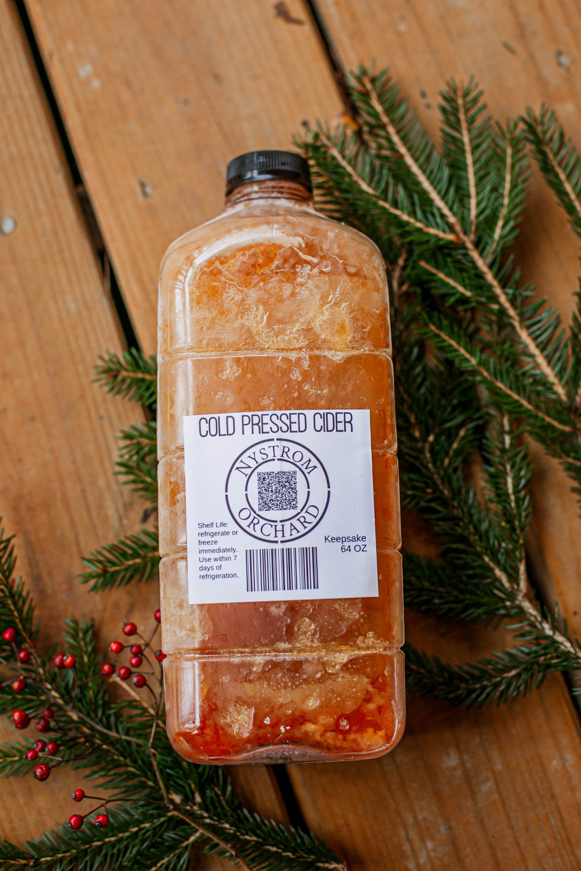 Bottle of Nystrom Orchard cold pressed cider on a wooden surface with greenery. Pressed in the kitchen of Nystrom Orchard in Worthington Minnesota.