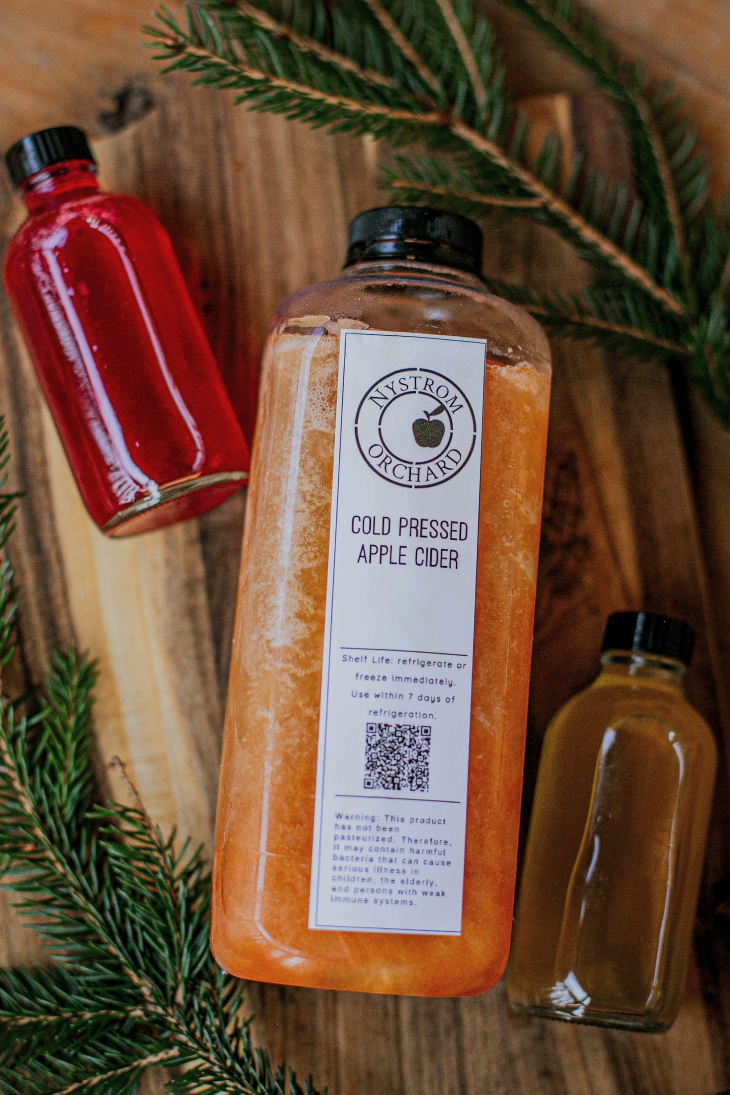 Bottle of cold pressed apple cider with a label on a wooden surface with two cider syrup bottles and greenery. Created in the kitchen of Nystrom Orchard in Worthington Minnesota. 