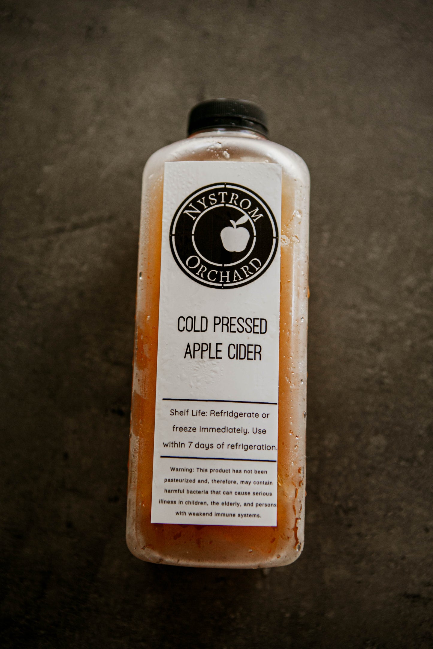 Bottle of Nystrom Orchard cold pressed cider on a black textured surface. Pressed in the kitchen at Nystrom Orchard in Worthington Minnesota. 