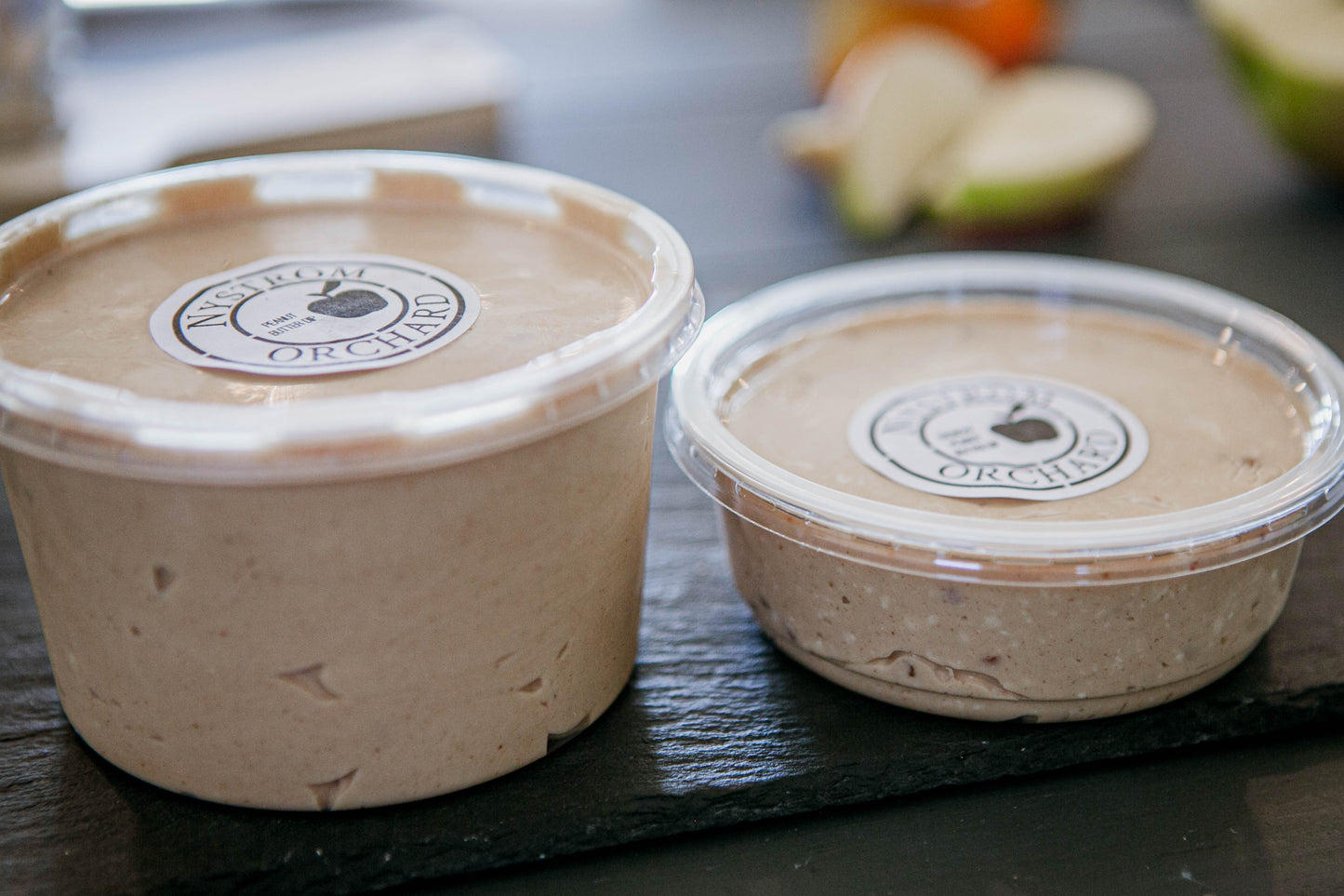 Two containers of peanut butter honey dip on a dark surface with blurred apples in the background. Made in the kitchen at Nystrom Orchard in Worthington Minnesota.