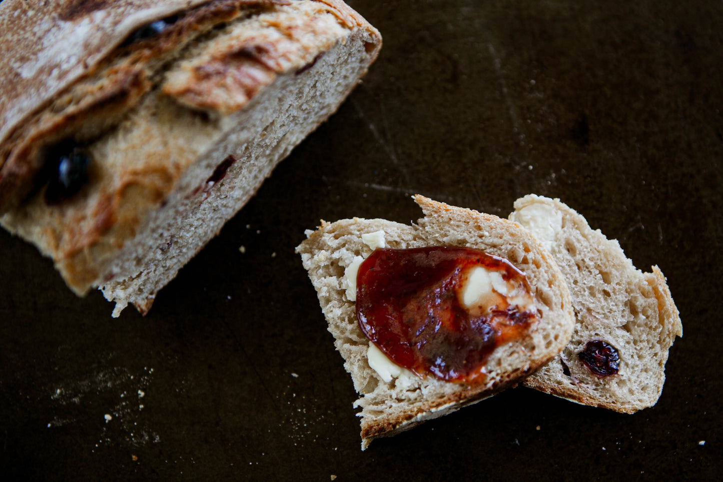 Loaf of Cranberry cream cheese sourdough bread cut with two slices in the center of the board. The one slice is spread with butter and jam on a dark surface. Baked in the kitchen at Nystrom Orchard in Worthington Minnesota.