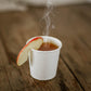 White paper cup with hot apple cider and an apple slice on a wooden surface. Cold pressed in the kitchen of Nystrom Orchard in Worthington Minnesota.