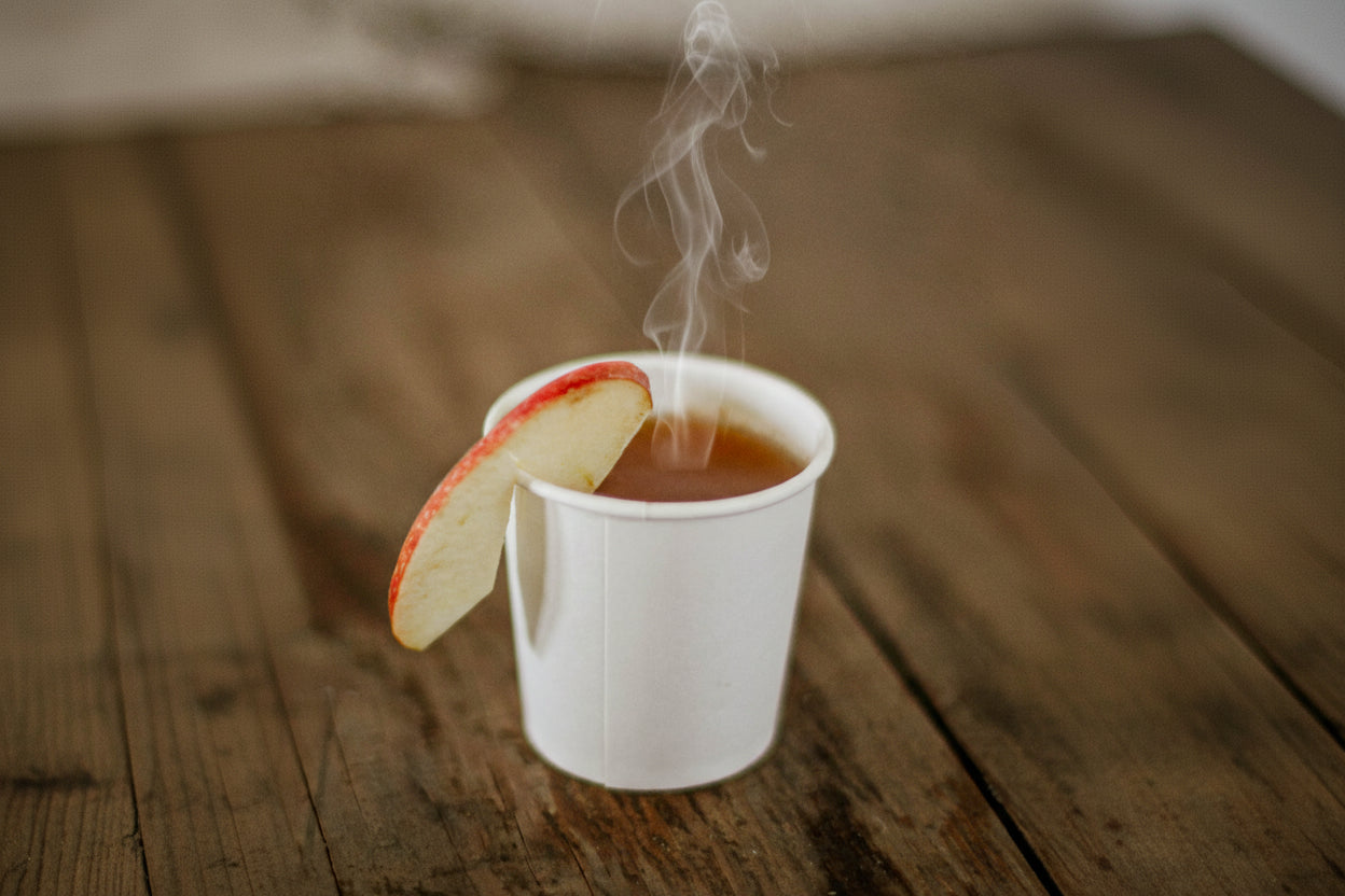 White paper cup with hot apple cider and an apple slice on a wooden surface. Cold pressed in the kitchen of Nystrom Orchard in Worthington Minnesota.