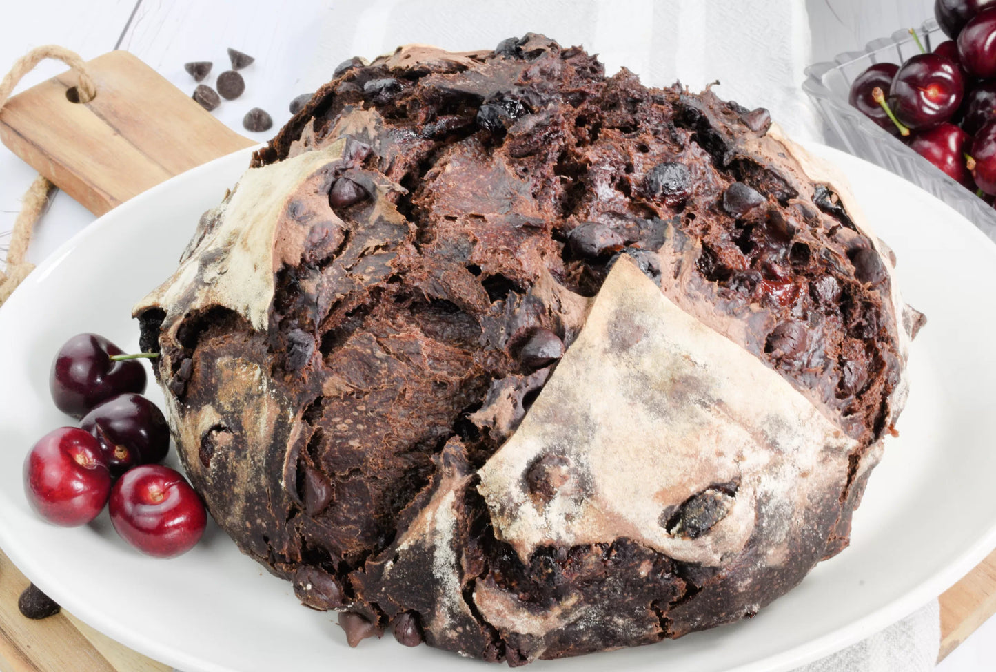A rustic loaf of chocolate cherry sourdough bread on a white plate on a cutting board with fresh sweet cherries beside. Baked in the kitchen of Nystrom Orchard in Worthington Minnesota.