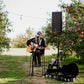 A person sitting in a high chair playing guitar with a mike and speaker in an orchard with string lights. There is a guitar case and a chair with some stuff on. Event in Nystrom Orchard in Worthington Minnesota. 