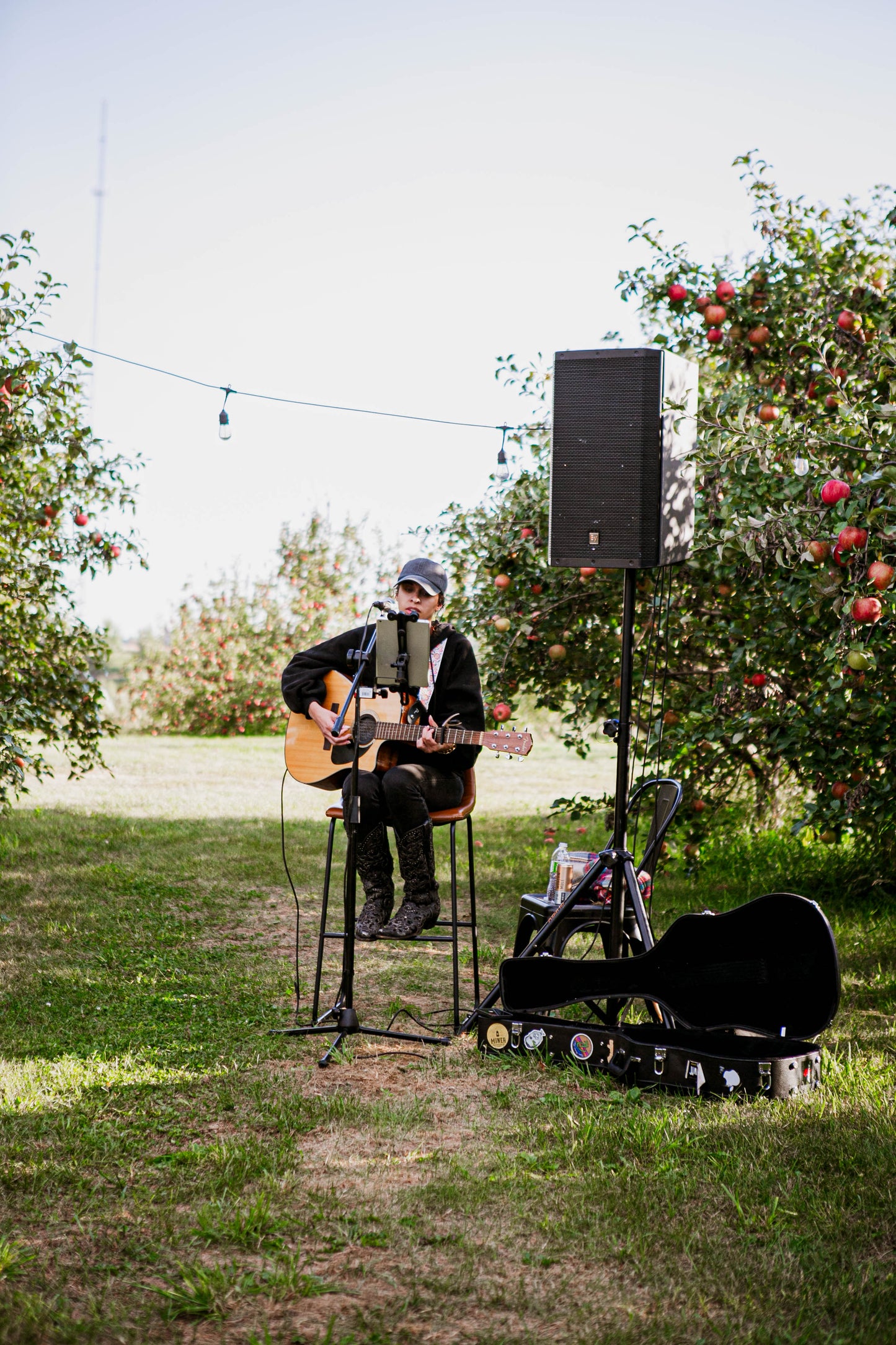 A person sitting in a high chair playing guitar with a mike and speaker in an orchard with string lights. There is a guitar case and a chair with some stuff on. Event in Nystrom Orchard in Worthington Minnesota. 