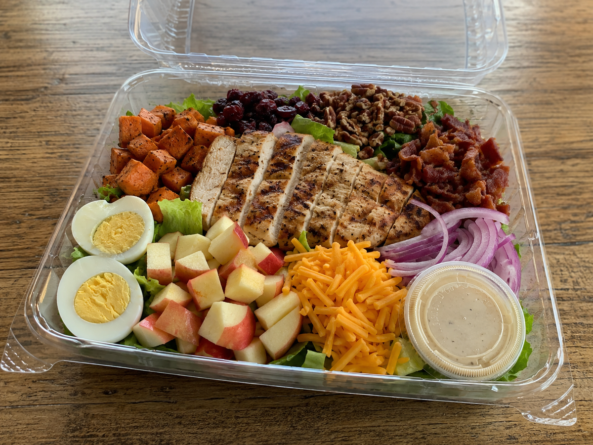 A clear plastic deli container filled with a mixed salad on a wooden table. The salad includes sliced grilled chicken breast, diced red apples, hard-boiled egg halves, shredded cheddar cheese, red onion, crumbled bacon, dark berries, sweet potato cubes, and a small clear cup of dressing. Made in the kitchen of Nystrom Orchard in Worthington Minnesota.