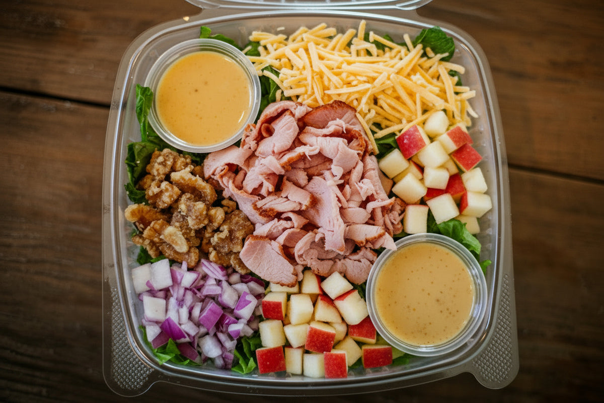 A clear plastic container filled with a prepared salad on a dark wooden table. The salad includes lettuce, shaved pork, diced apples, chopped red onion, cranberries, nuts, and two small cups of honey mustard dressing. Made in the kitchen of Nystrom Orchard in Worthington Minnesota.