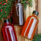Three glass bottles of flavored apple cider syrup on a wooden background with greenery and red berries. Made by the kitchen at Nystrom Orchard in Worthington Minnesota.