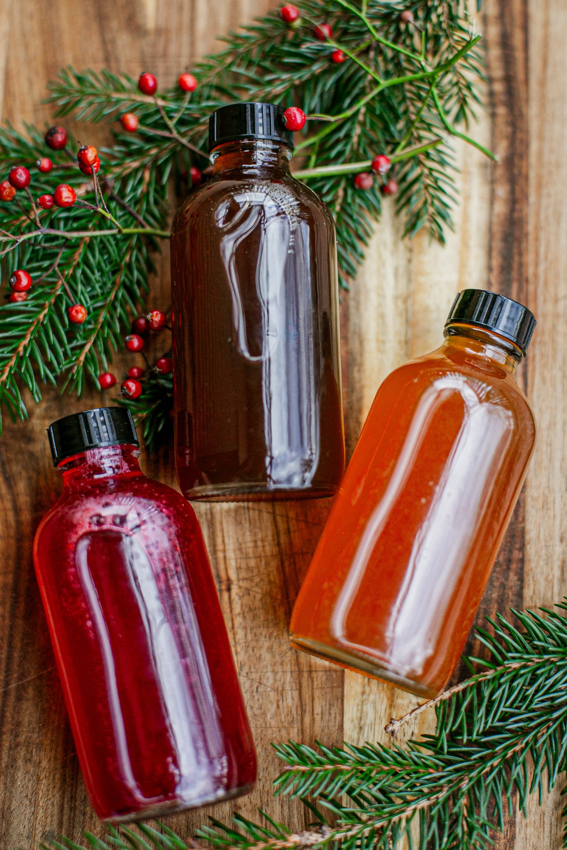 Three glass bottles of flavored apple cider syrup on a wooden background with greenery and red berries. Made by the kitchen at Nystrom Orchard in Worthington Minnesota.