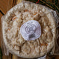Frozen apple pie in plastic with a sticker. The pie is on a wooden cutting board with greenery around. Made in the kitchen of Nystrom Orchard in Worthington Minnesota.