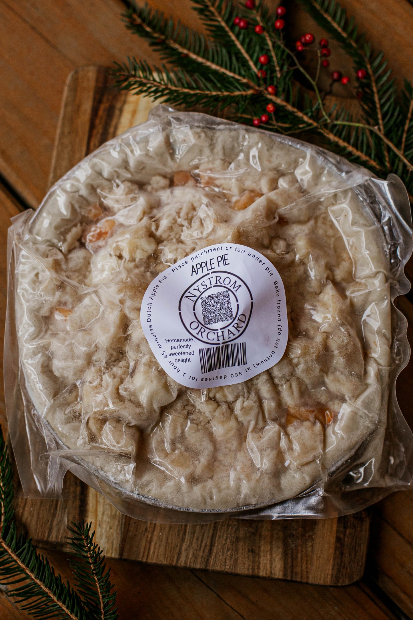 Frozen apple pie in plastic with a sticker. The pie is on a wooden cutting board with greenery around. Made in the kitchen of Nystrom Orchard in Worthington Minnesota.