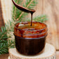 Glass jar with fudge syrup and a spoon drizzling syrup back into the jar on a wooden surface with greenery. Made in the kitchen at Nystrom Orchard in Worthington Minnesota.