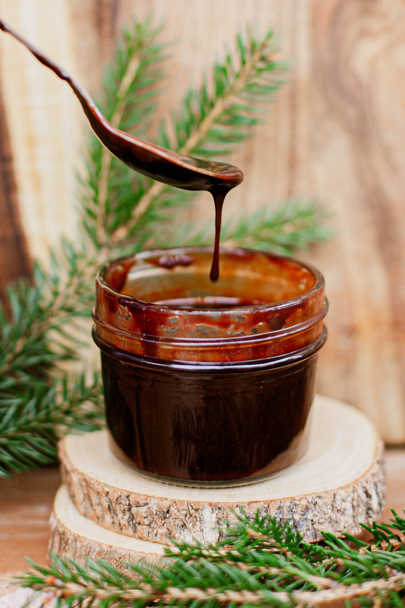 Glass jar with fudge syrup and a spoon drizzling syrup back into the jar on a wooden surface with greenery. Made in the kitchen at Nystrom Orchard in Worthington Minnesota.