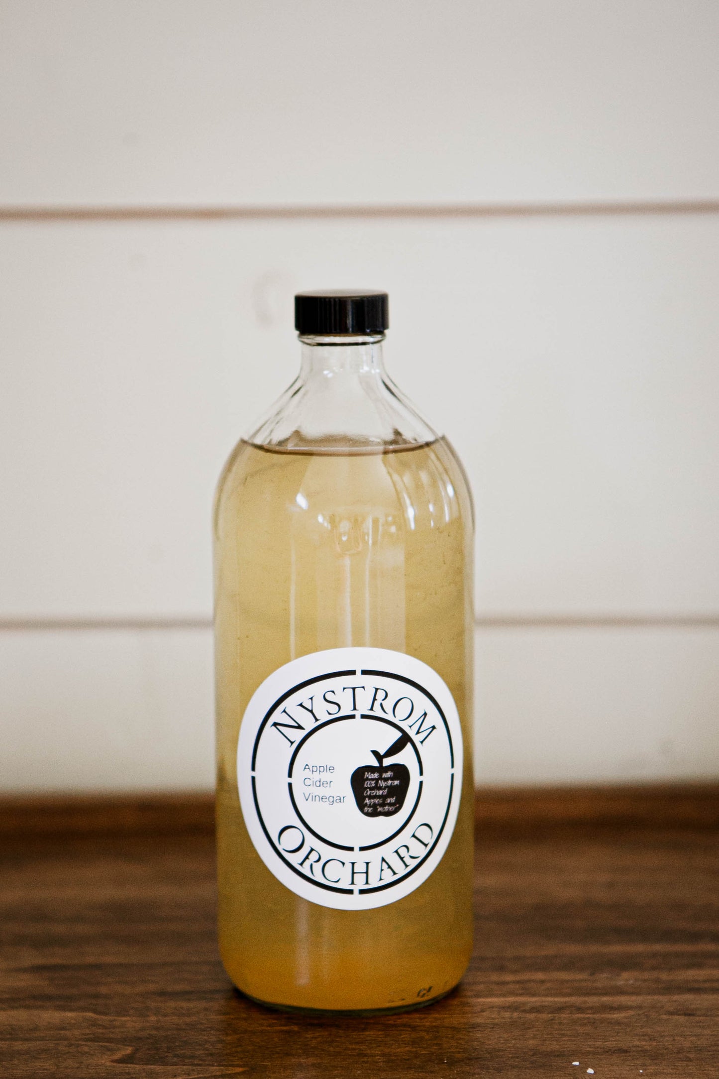 Glass bottle of house made apple cider vinegar on a wooden surface with a white background. Made in the kitchen of Nystrom Orchard in Worthington Minnesota.