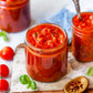 Jars of homemade marinara sauce on a wooden board, surrounded by vine-ripened tomatoes and a spoon with sauce on it, indicating freshness and homemade quality. Made in the kitchen of Nystrom Orchard in Worthington Minnesota.