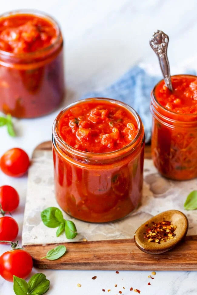 Jars of homemade marinara sauce on a wooden board, surrounded by vine-ripened tomatoes and a spoon with sauce on it, indicating freshness and homemade quality. Made in the kitchen of Nystrom Orchard in Worthington Minnesota.