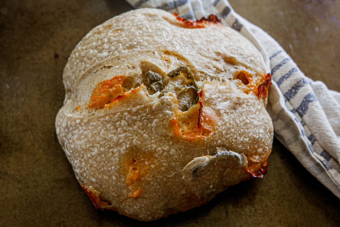Rustic loaf of sourdough bread with cheddar and jalapenos on a dark surface with a blue and white cloth. Baked in the kitchen at Nystrom Orchard in Worthington Minnesota. 