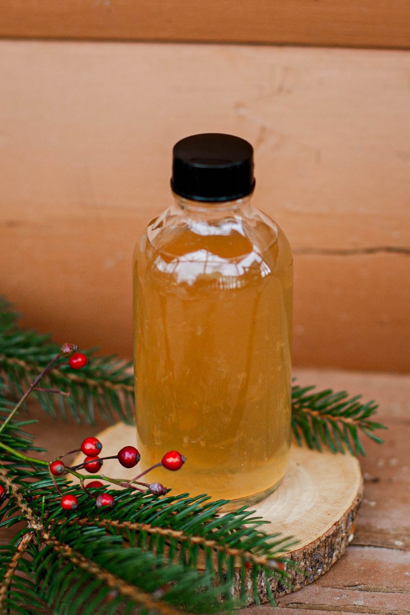 Glass bottle with lemon ginger syrup and a black cap on a wooden surface with a wooden background, greenery and red berries. Made in the kitchen at Nystrom Orchard in Worthington Minnesota.