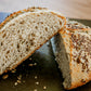 Two halves of a loaf of rustic hearty sourdough bread with seeds on a dark surface with seeds scattered around them. Baked in the kitchen at Nystrom Orchard in Worthington Minnesota.