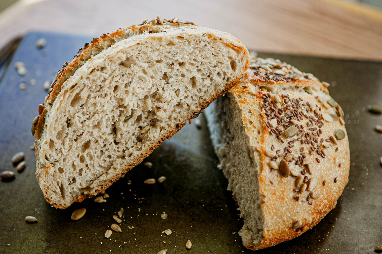 Two halves of a loaf of rustic hearty sourdough bread with seeds on a dark surface with seeds scattered around them. Baked in the kitchen at Nystrom Orchard in Worthington Minnesota.