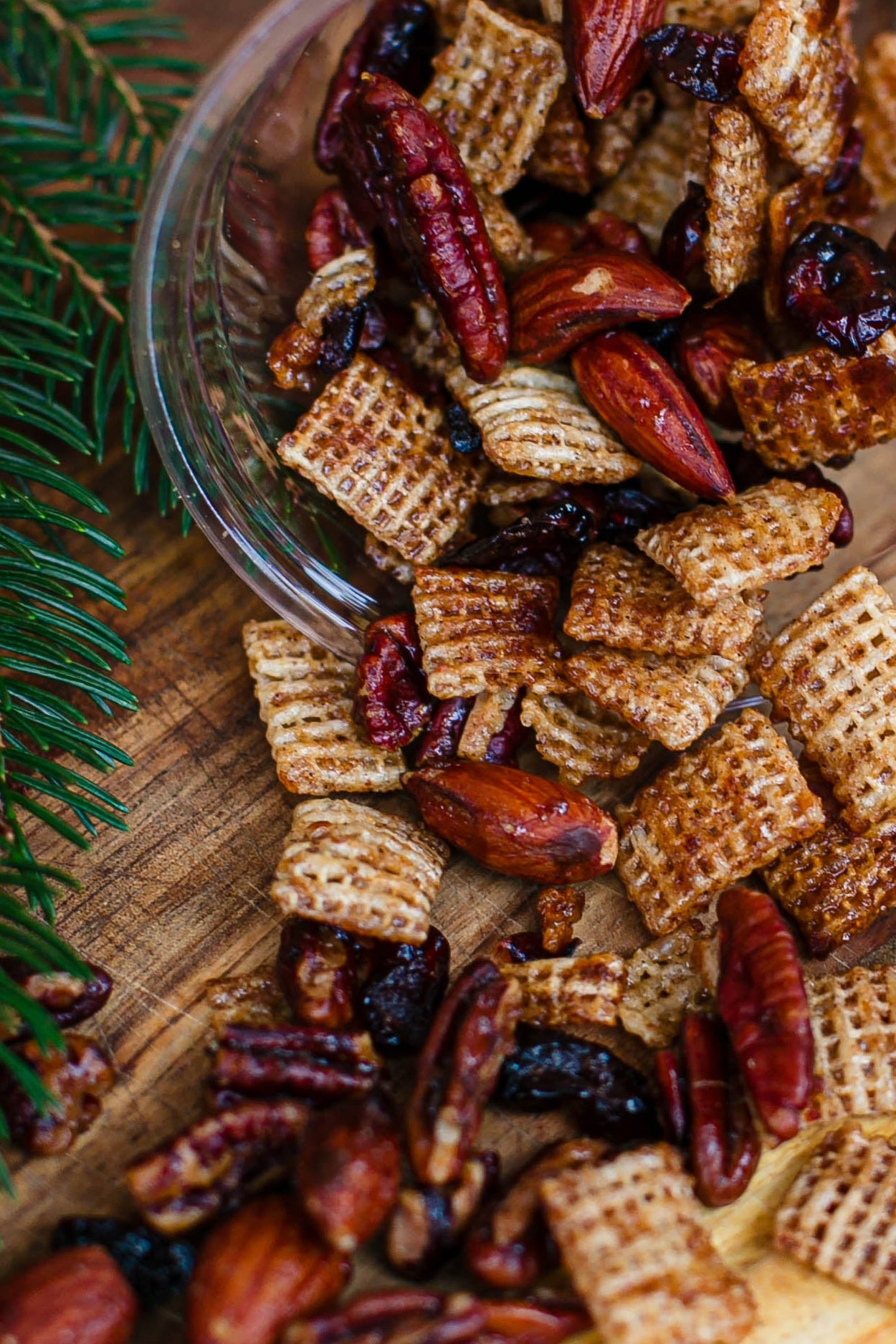 Nuts, cranberries, and rice chex mixed together in a snack mix spilling out of a cup onto a wooden surface with greenery. Made by the kitchen at Nystrom Orchard in Worthington Minnesota.