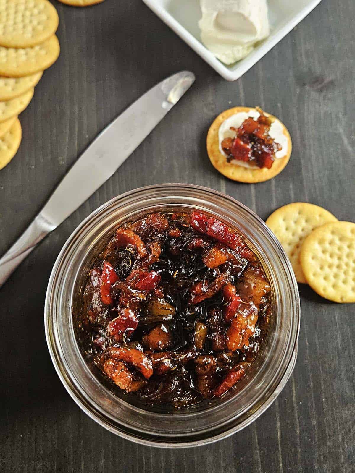 Jar of maple onion bacon jam surrounded by crackers and a cream cheese along with a knife on a dark surface. Made in the kitchen at Nystrom Orchard in Worthington Minnesota