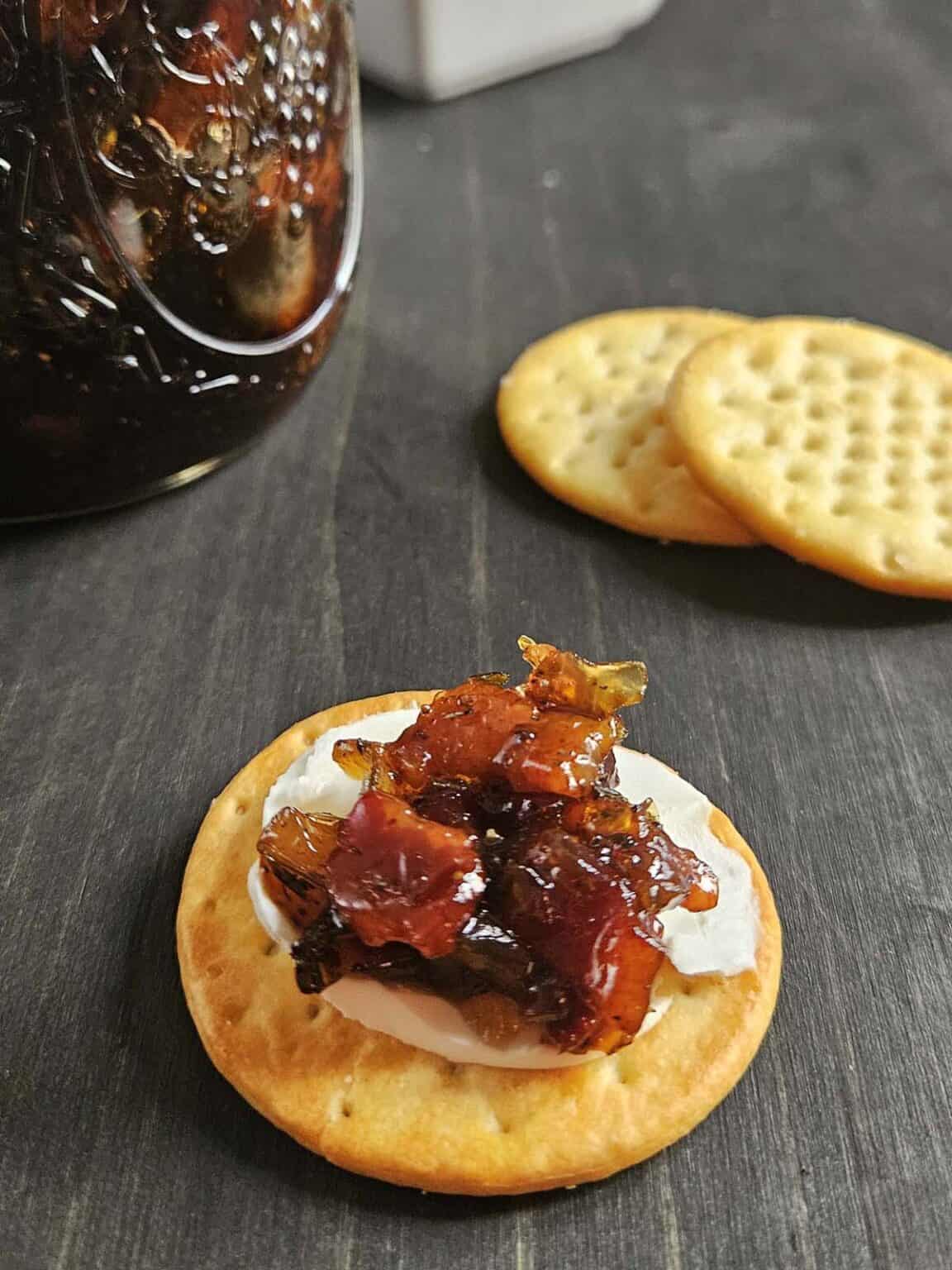 Cracker with cream cheese and maple bacon onion jam on a dark surface. Made in the kitchen at Nystrom Orchard in Worthington Minnesota.