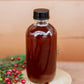 Glass bottle of maple cider syrup with a black cap with a wooden background and greenery and red berries. Made in the kitchen at Nystrom Orchard in Worthington Minnesota. 