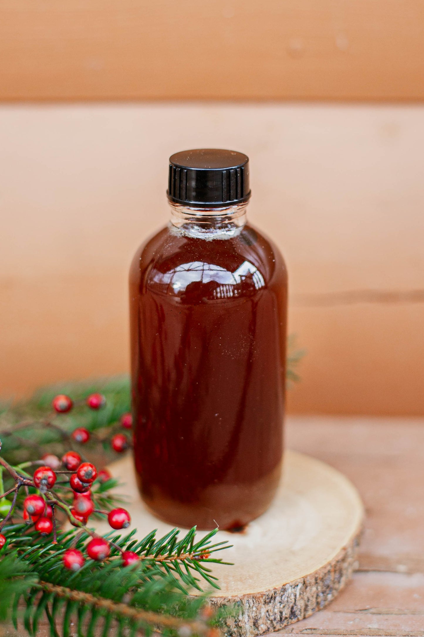 Glass bottle of maple cider syrup with a black cap with a wooden background and greenery and red berries. Made in the kitchen at Nystrom Orchard in Worthington Minnesota. 