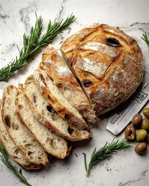 Sliced rustic artisan bread with a flour-dusted crust and dark olive pits, arranged on a light marble surface with rosemary sprigs, olives, on a paper. Baked in the kitchen of Nystrom Orchard in Worthington Minnesota.