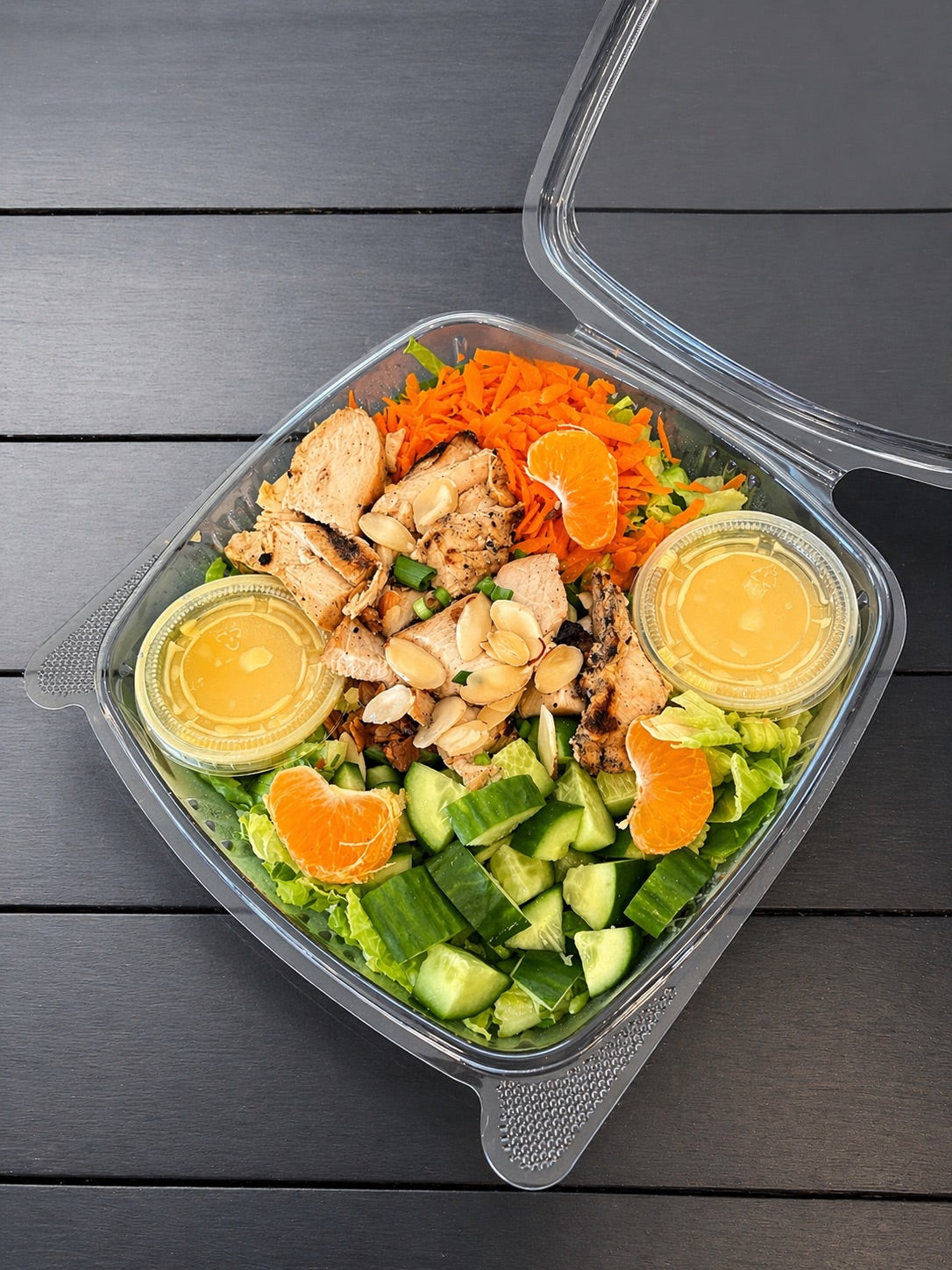 Salad with chicken, carrots, oranges, cucumbers, and citrus dressing in a small container in a clear container on a wooden surface. Made fresh in the kitchen at Nystrom Orchard in Worthington Minnesota.