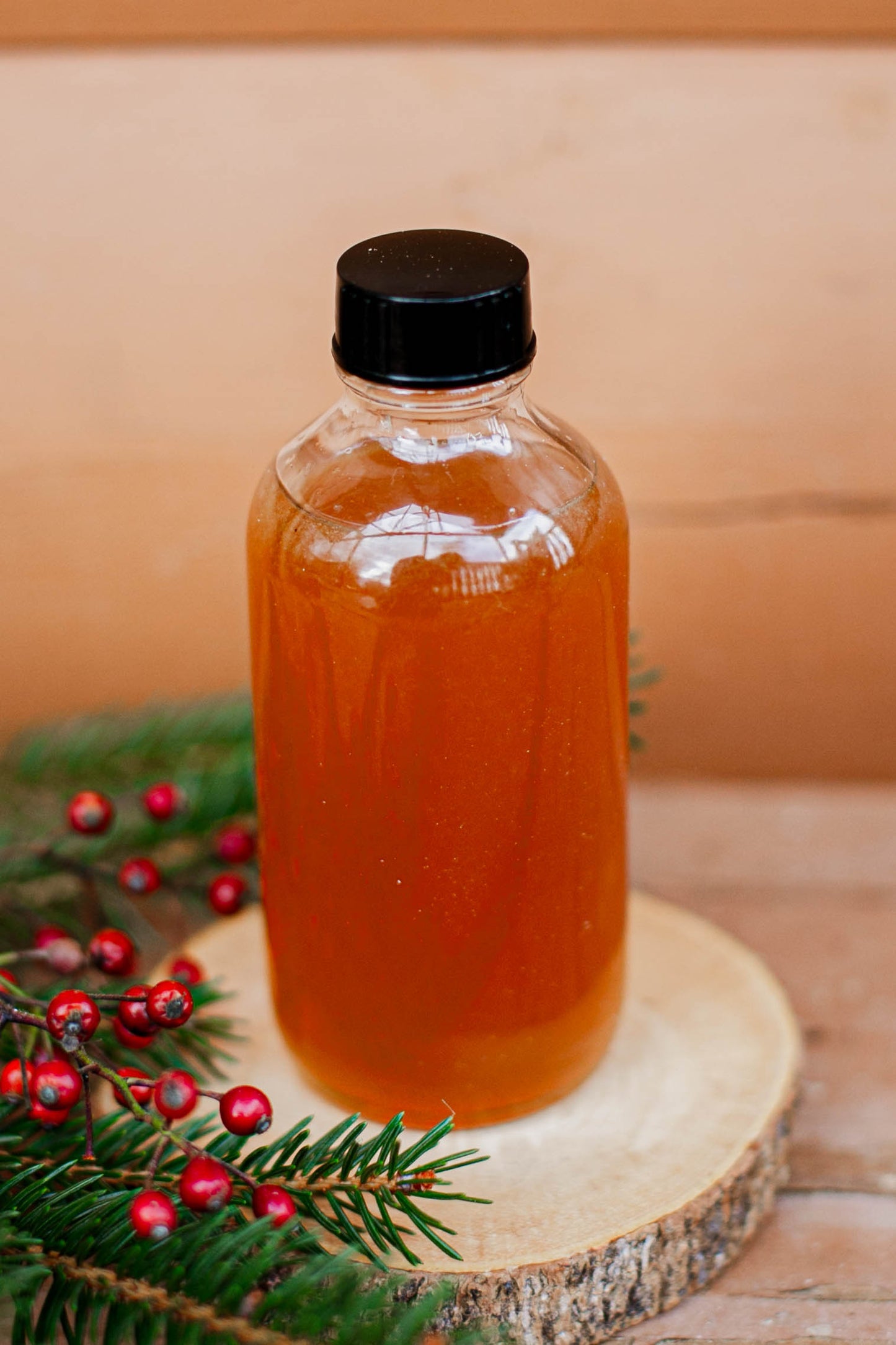 Glass bottle with Nystrom orchard signature syrup with a black cap on a wooden surface with greenery and red berries. Made in the kitchen at Nystrom Orchard in Worthington Minnesota.