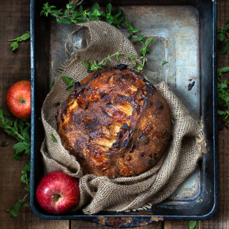 Rustic loaf of sourdough bread filled with apples, onions, and sage on a rustic metal tray with burlap, greenery and apples. Baked in the kitchen at Nystrom Orchard in Worthington Minnesota. 