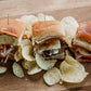 Three sliders on buns with various fillings on a wooden board with potato chips. Made in the kitchen of Nystrom Orchard in Worthington Minnesota.