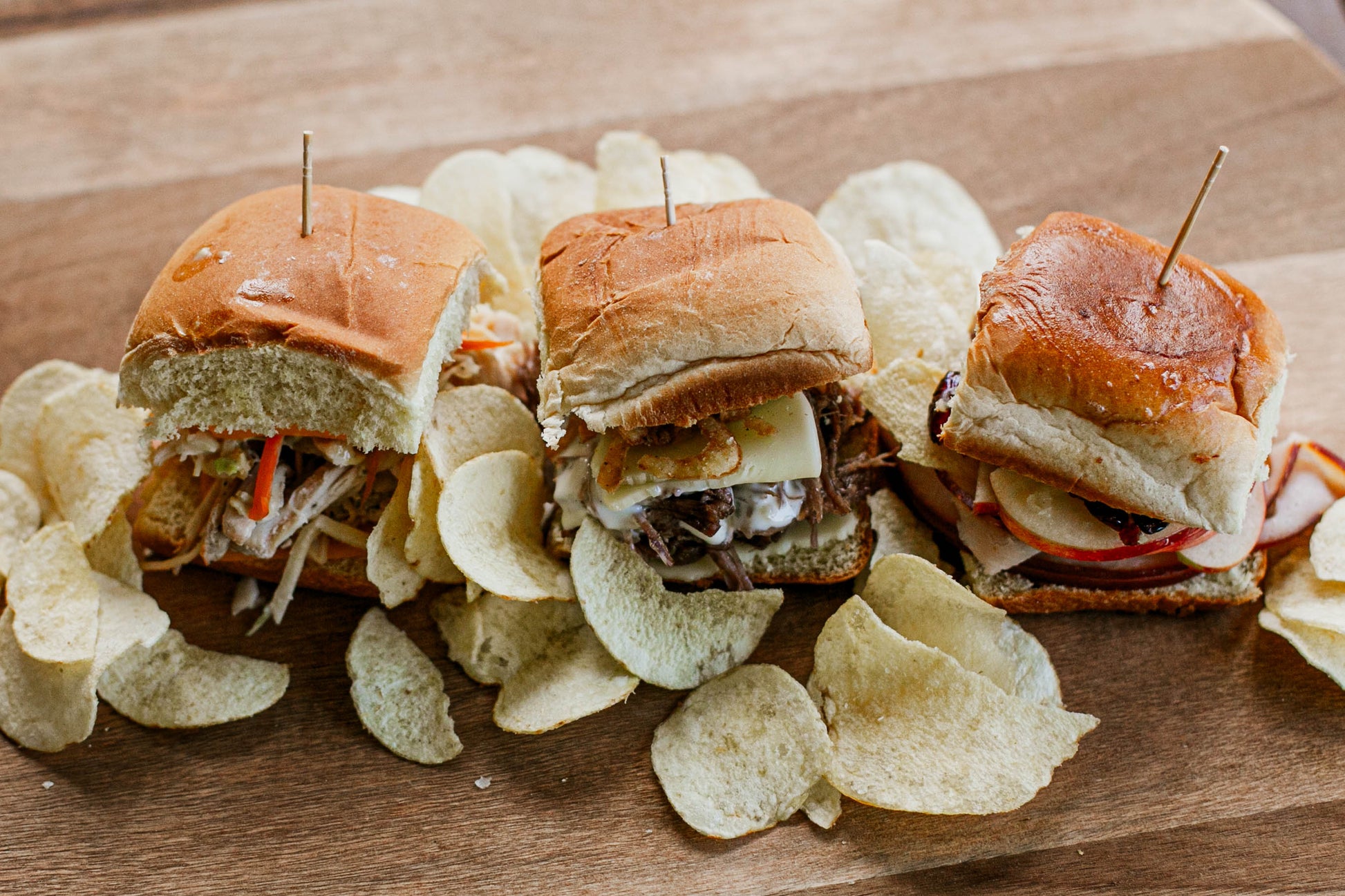 Three sliders on buns with various fillings on a wooden board with potato chips. Made in the kitchen of Nystrom Orchard in Worthington Minnesota.