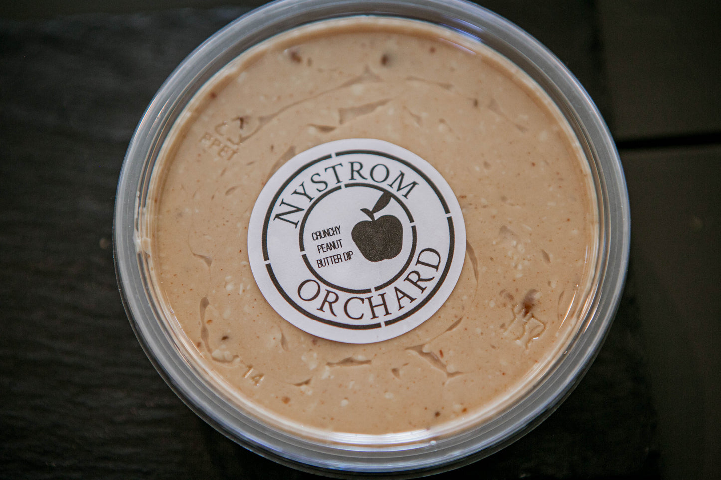 A plastic container of peanut butter cream cheese apple dip with a label that reads 'Nystrom Orchard' and has an image of an apple. Made in the kitchen of Nystrom Orchard in Worthington Minnesota.