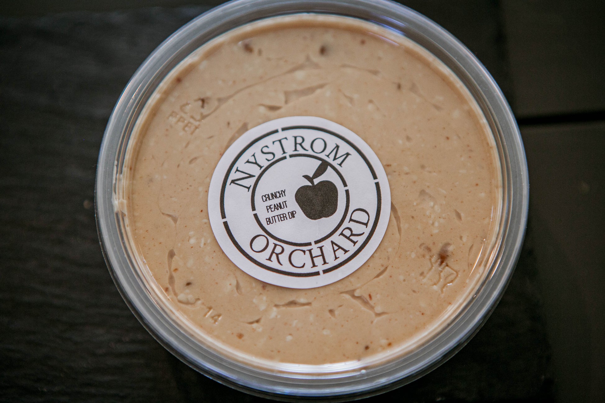 A plastic container of peanut butter cream cheese apple dip with a label that reads 'Nystrom Orchard' and has an image of an apple. Made in the kitchen of Nystrom Orchard in Worthington Minnesota.