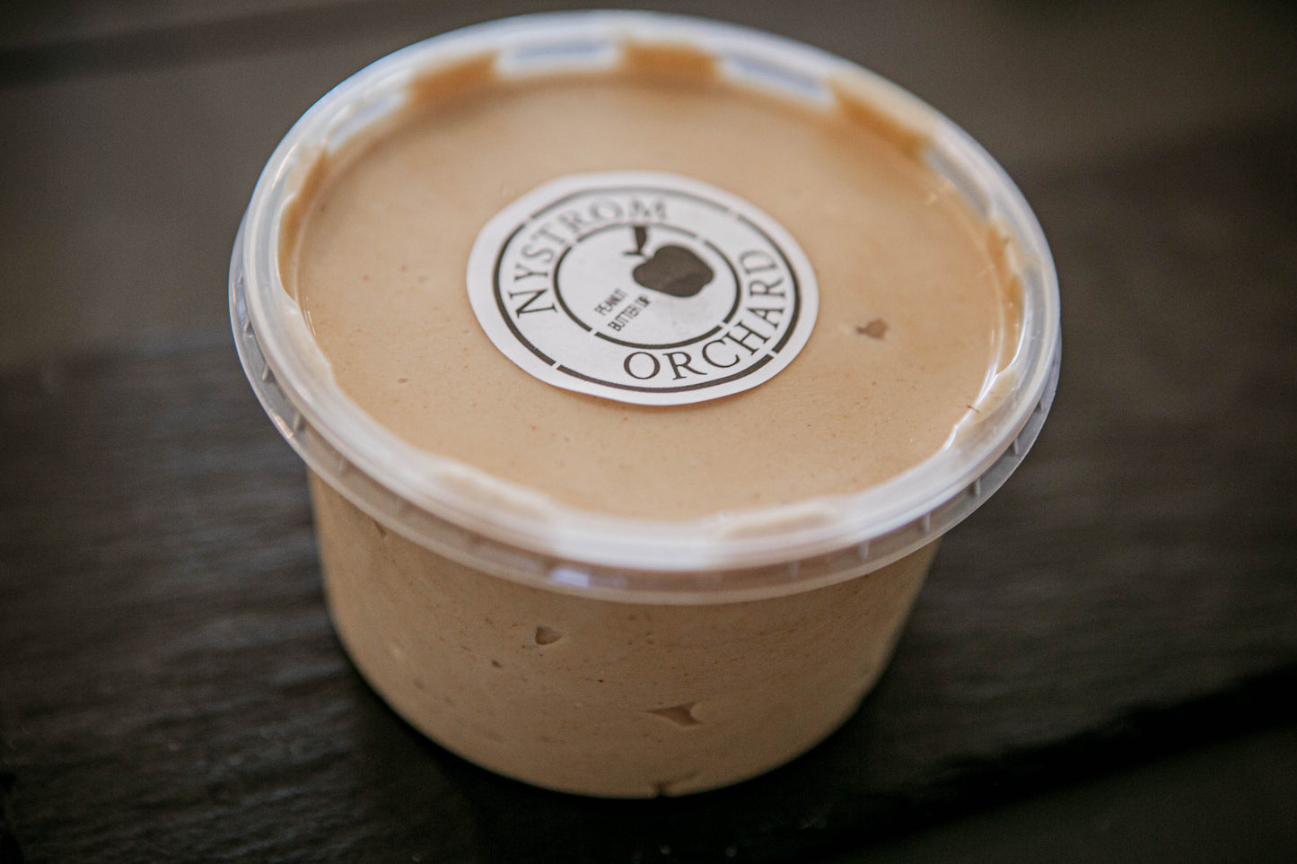 A plastic container of peanut butter cream cheese apple dip with a label that reads 'Nystrom Orchard' and has an image of an apple on a dark surface. Made in the kitchen of Nystrom Orchard in Worthington Minnesota. 