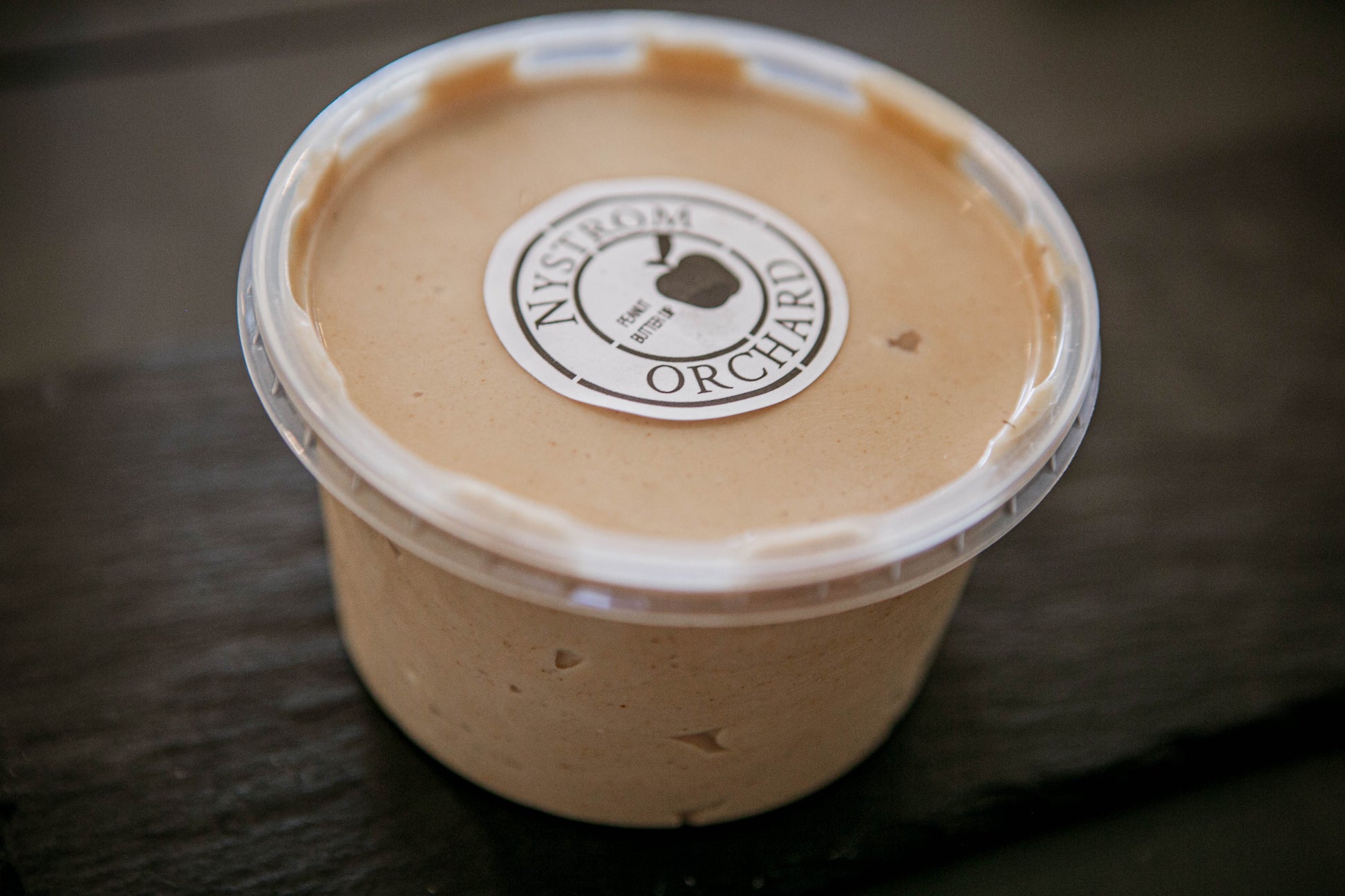 A plastic container of peanut butter cream cheese apple dip with a label that reads 'Nystrom Orchard' and has an image of an apple on a dark surface. Made in the kitchen of Nystrom Orchard in Worthington Minnesota. 