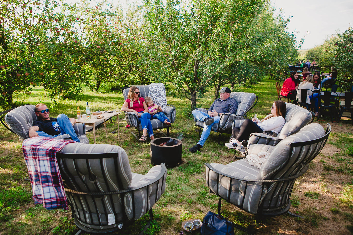 A group of people sitting around the campfire on soft chairs with apple trees in the background and people eating at table and chairs. Event hosted by Nystrom Orchard in Worthington Minnesota.