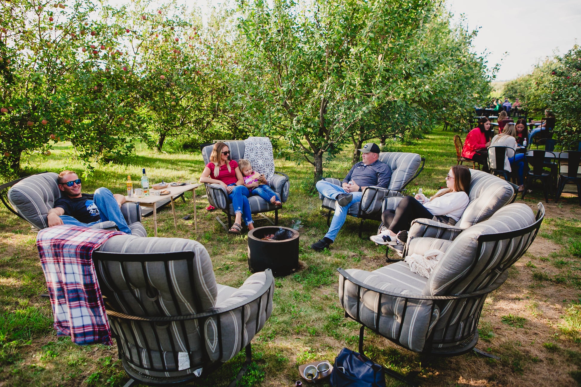 A group of people sitting around the campfire on soft chairs with apple trees in the background and people eating at table and chairs. Event hosted by Nystrom Orchard in Worthington Minnesota.