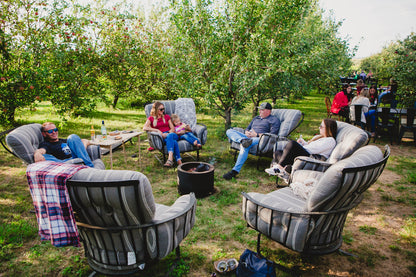A group of people sitting around the campfire on soft chairs with apple trees in the background and people eating at table and chairs. Event hosted by Nystrom Orchard in Worthington Minnesota.