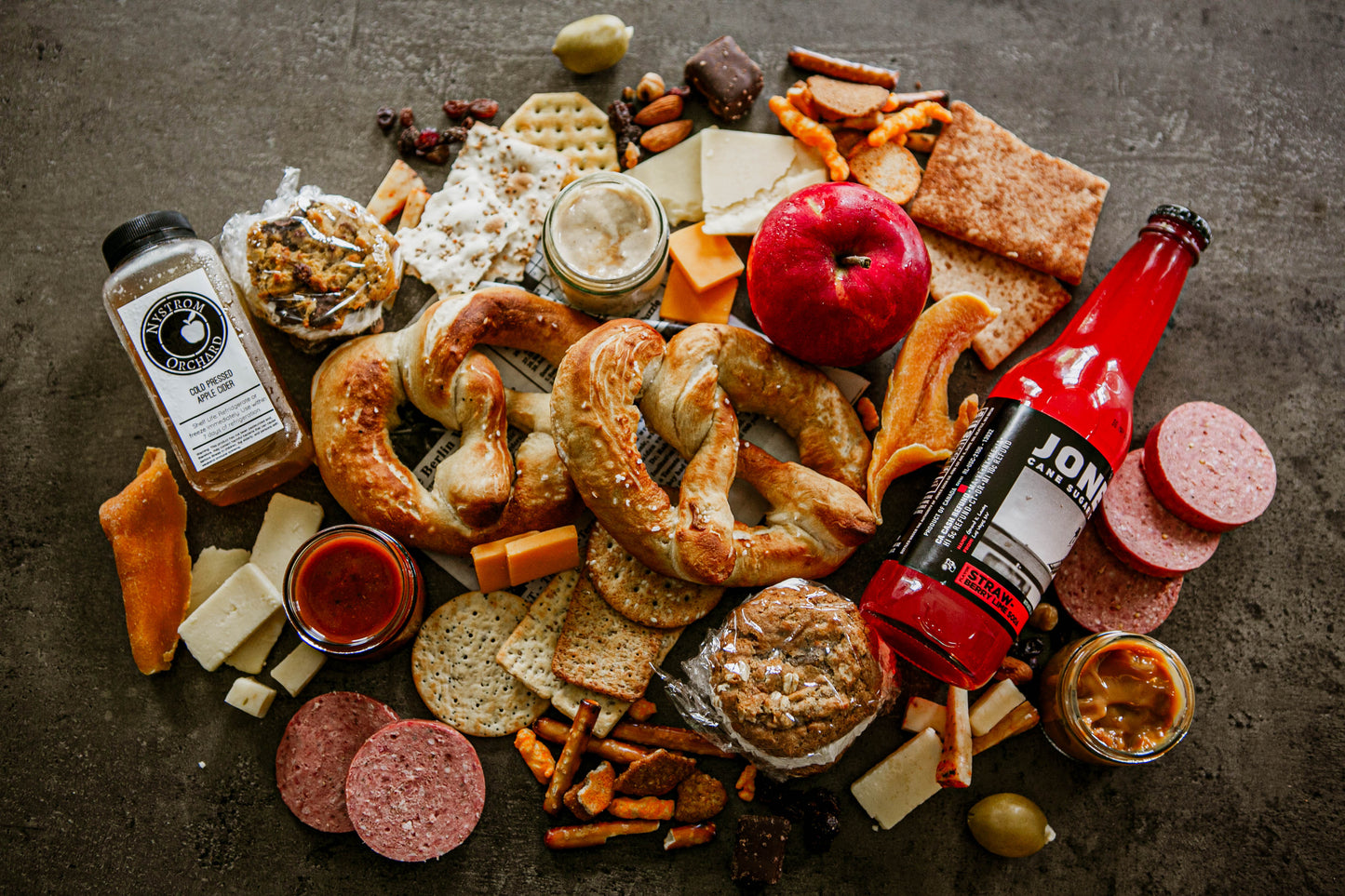 A picnic spread of soft pretzels misc. crackers, whoopie pies, dips, cheese, dried fruits, apples and drinks on a dark surface. Created in the kitchen of Nystrom Orchard in Worthington Minnesota.