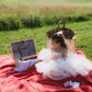 Child in a white dress sitting on a red blanket with a picnic setup in a grassy field. Created in the kitchen of Nystrom Orchard in Worthington Minnesota.